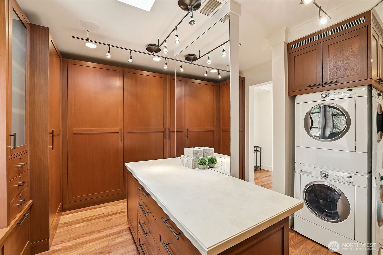 This functional laundry room features a stacked washer and dryer unit set against warm-toned cabinetry that provides ample storage. A central island with a light-colored countertop offers a convenient workspace, while track lighting mounted on the ceiling illuminates the area. The room is finished with light wood flooring and a large mirror that enhances the sense of space.