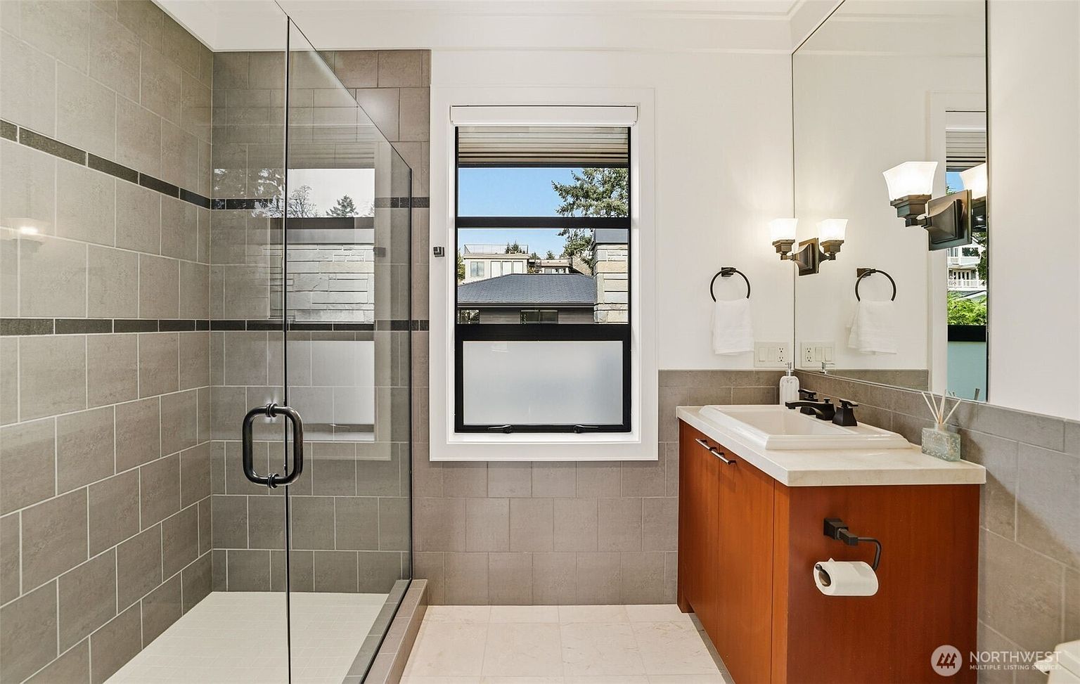 This modern bathroom features a glass-enclosed walk-in shower with gray tiled walls and a contrasting dark accent stripe. A wooden vanity with a light-colored countertop and a rectangular sink sits beneath a large mirror, complemented by dual wall-mounted light fixtures. A window with a frosted lower pane provides natural light, while a toilet paper holder is conveniently mounted to the side of the vanity.