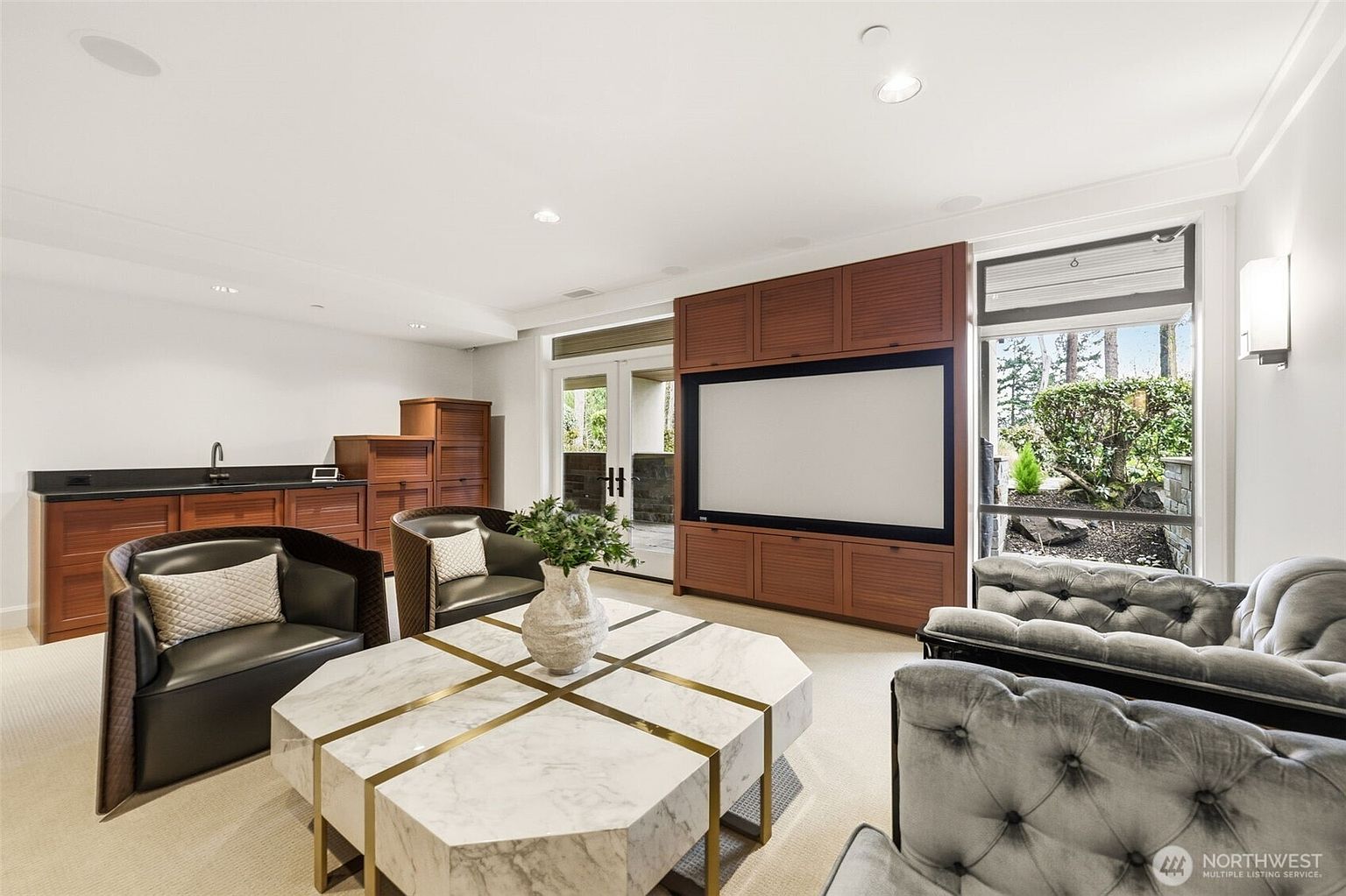 This sophisticated media room features a large projection screen integrated into a custom wood cabinetry wall, flanked by a wet bar area on the left. The seating arrangement includes two dark leather armchairs and a tufted grey sofa centered around a large, geometric stone-topped coffee table. Large windows provide a view of the outdoor greenery, while recessed lighting and a neutral color palette create a refined, comfortable atmosphere.