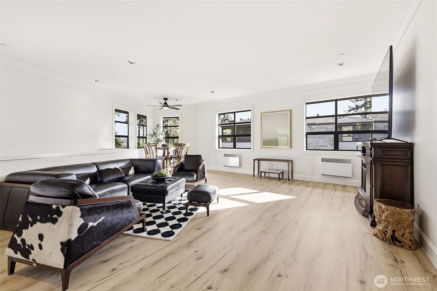 This spacious, open-concept living area features light wood-look flooring and a bright, neutral color palette. The room is furnished with a dark leather sectional, a matching ottoman, and a unique cowhide-patterned armchair, all anchored by a bold black-and-white geometric rug. Large windows allow ample natural light to fill the space, which also includes a dining area in the background and a traditional-style media console with a television.