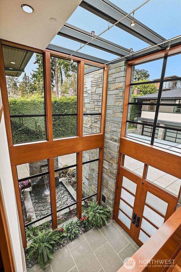 This dramatic entryway features a soaring glass-paneled roof and expansive windows that flood the space with natural light. A striking stone-clad wall serves as a focal point, complemented by a serene indoor water feature and lush greenery at the base. The combination of warm wood framing, stone tile flooring, and modern architectural lines creates a sophisticated and welcoming atmosphere.