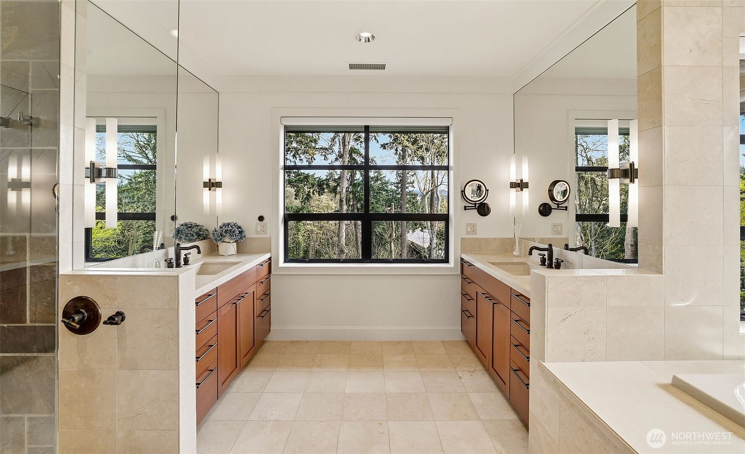This spacious primary bathroom features a symmetrical layout with dual vanities flanking a central window that offers a view of the surrounding trees. The room is finished with light-toned stone countertops and flooring, complemented by warm-toned wood cabinetry and large mirrors. The perspective is centered, highlighting the clean, balanced design and the bright, airy atmosphere of the space.