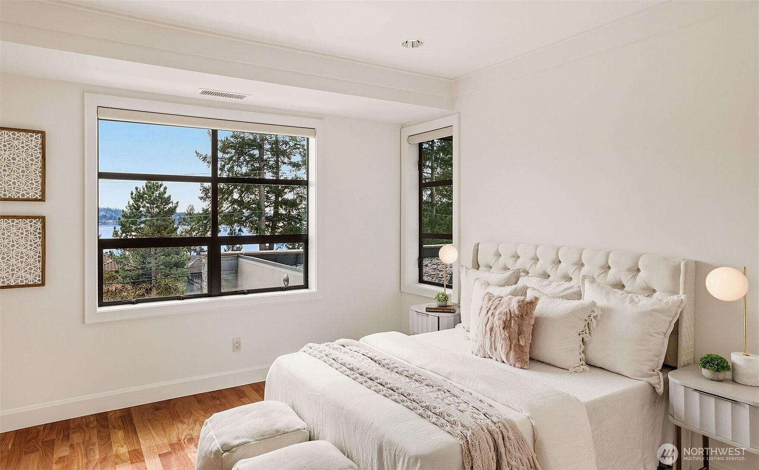 This bright and airy primary bedroom features a tufted, light-colored headboard as the focal point, complemented by neutral bedding and a textured throw blanket. Large windows provide ample natural light and a scenic view of the outdoors, while two modern globe-style lamps sit on matching nightstands. The room is finished with warm-toned wood flooring and minimalist wall art, creating a serene and inviting atmosphere.