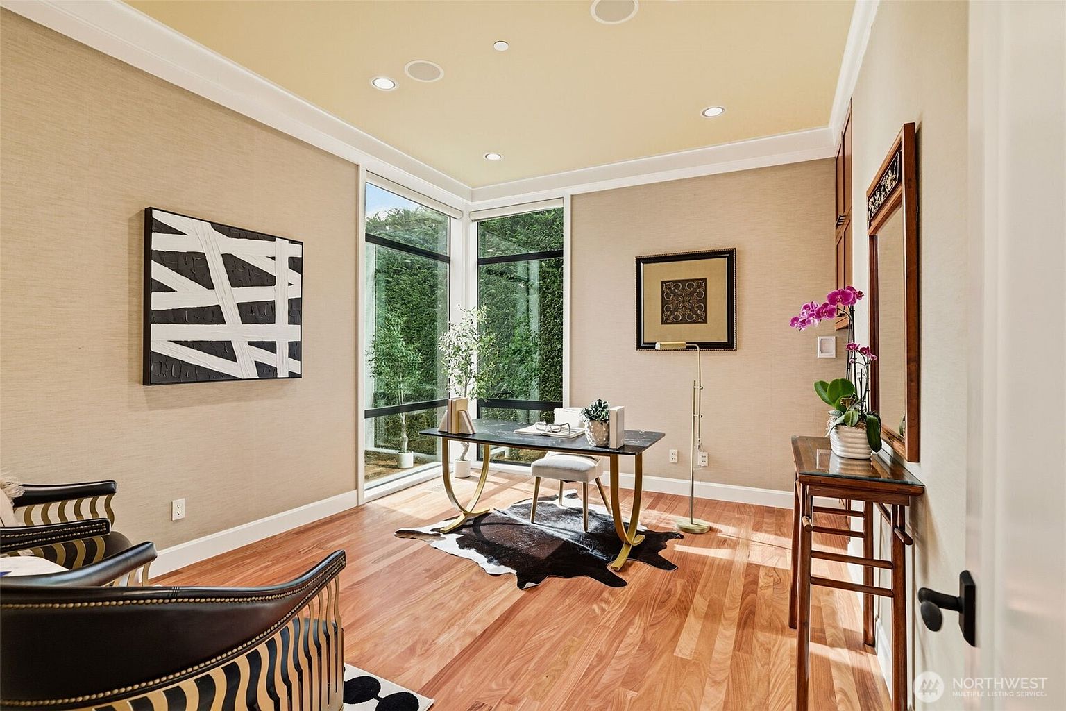 This sophisticated home office features warm, textured wall coverings and rich wood flooring, creating a professional yet inviting atmosphere. A sleek desk with a dark top and gold-toned legs sits atop a cowhide rug, positioned to take advantage of the natural light from the large windows that look out onto greenery. The room is accented by a piece of abstract wall art, a tall mirror over a console table with a potted orchid, and a pair of leather chairs in the foreground.