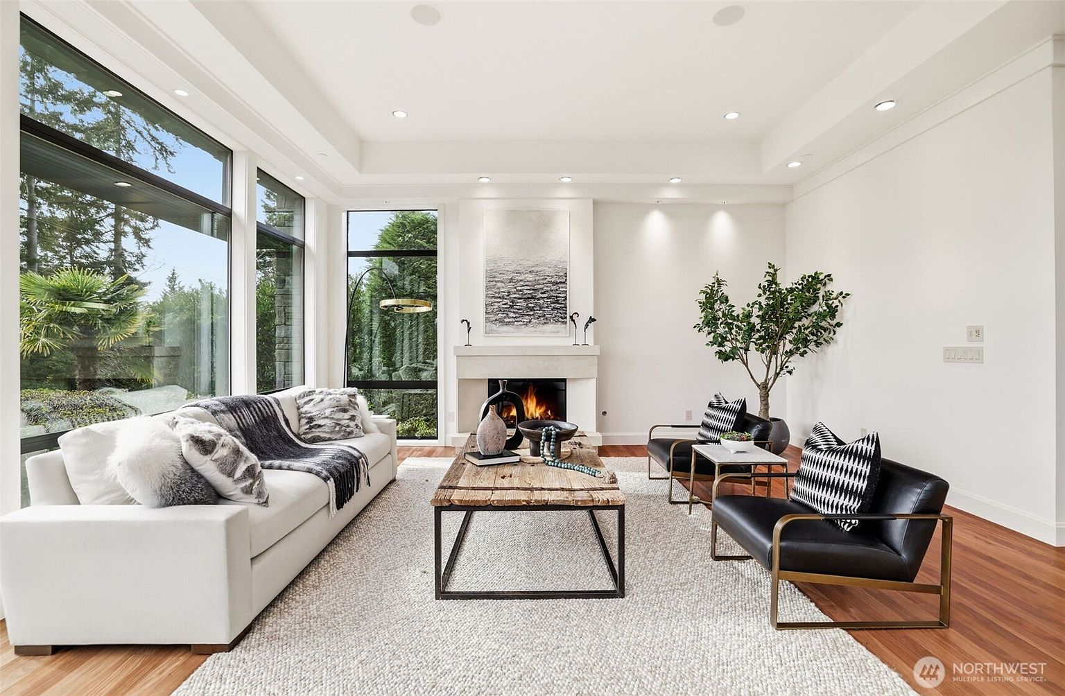 This bright and airy living room features a contemporary design with a neutral color palette and warm wood flooring. A large white sofa sits opposite two black leather armchairs, centered around a rustic wooden coffee table and a fireplace with a tall, minimalist surround. Expansive windows along the left wall provide abundant natural light and views of the surrounding greenery, creating a seamless connection between the indoor and outdoor spaces.