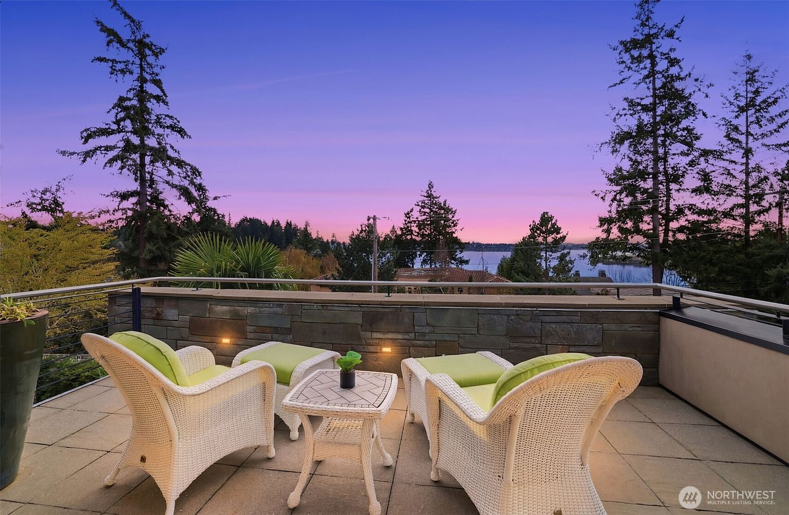 This elevated balcony offers a serene outdoor seating area with two white wicker chairs featuring lime green cushions, arranged around a small, patterned-tile bistro table. The space is framed by a stone-clad wall and a sleek metal railing, providing an unobstructed view of a tranquil lake at sunset. The soft, ambient lighting on the balcony complements the vibrant purple and orange hues of the twilight sky, creating a peaceful and inviting atmosphere.