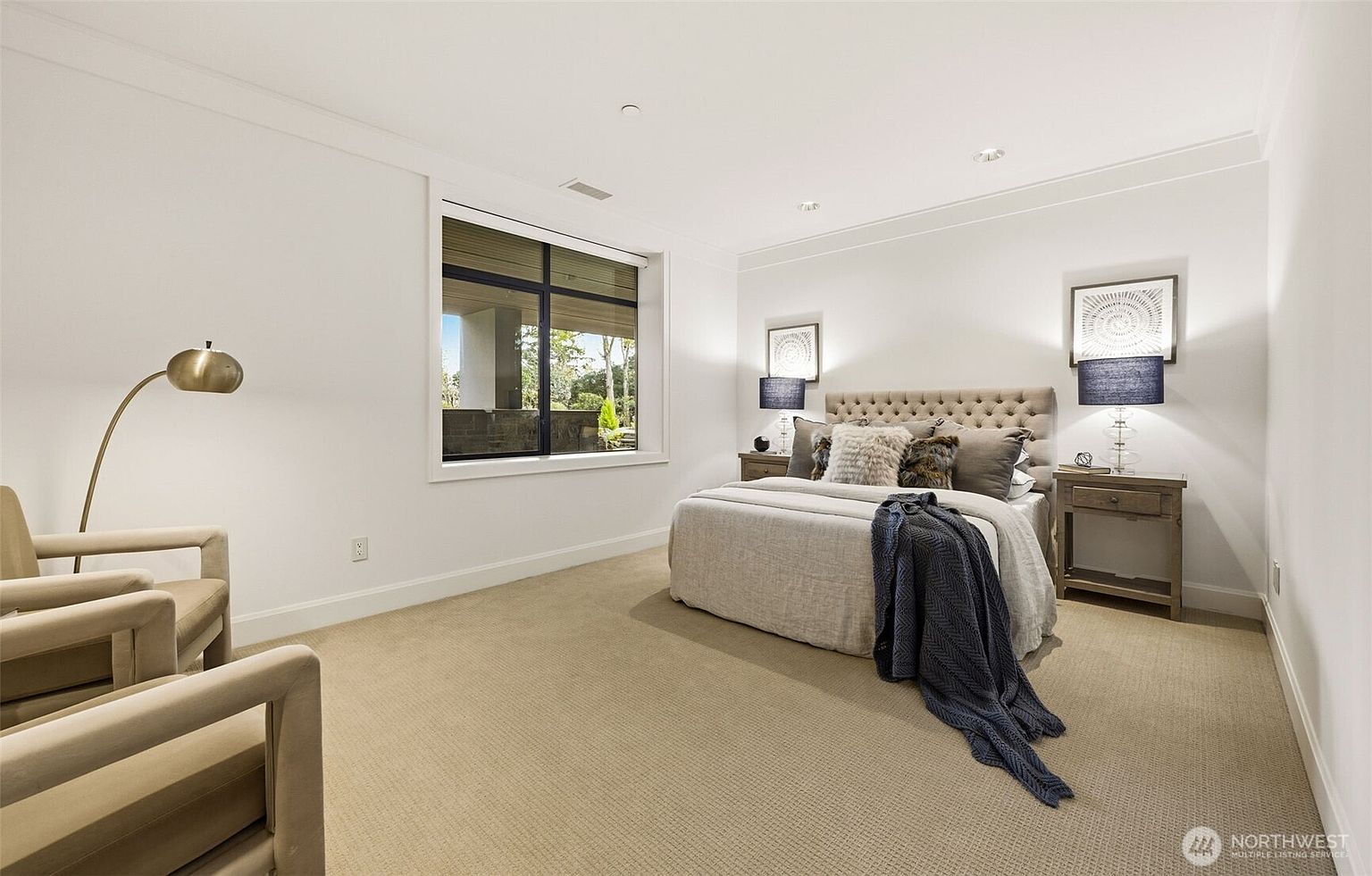 This bright and neutral bedroom features a plush, tufted headboard centered against a wall with matching nightstands and lamps. A large window provides natural light and a view of the outdoor greenery, while two comfortable armchairs and a floor lamp create a cozy seating area in the foreground. The room is finished with light-toned carpeting and recessed lighting, offering a clean and inviting atmosphere.