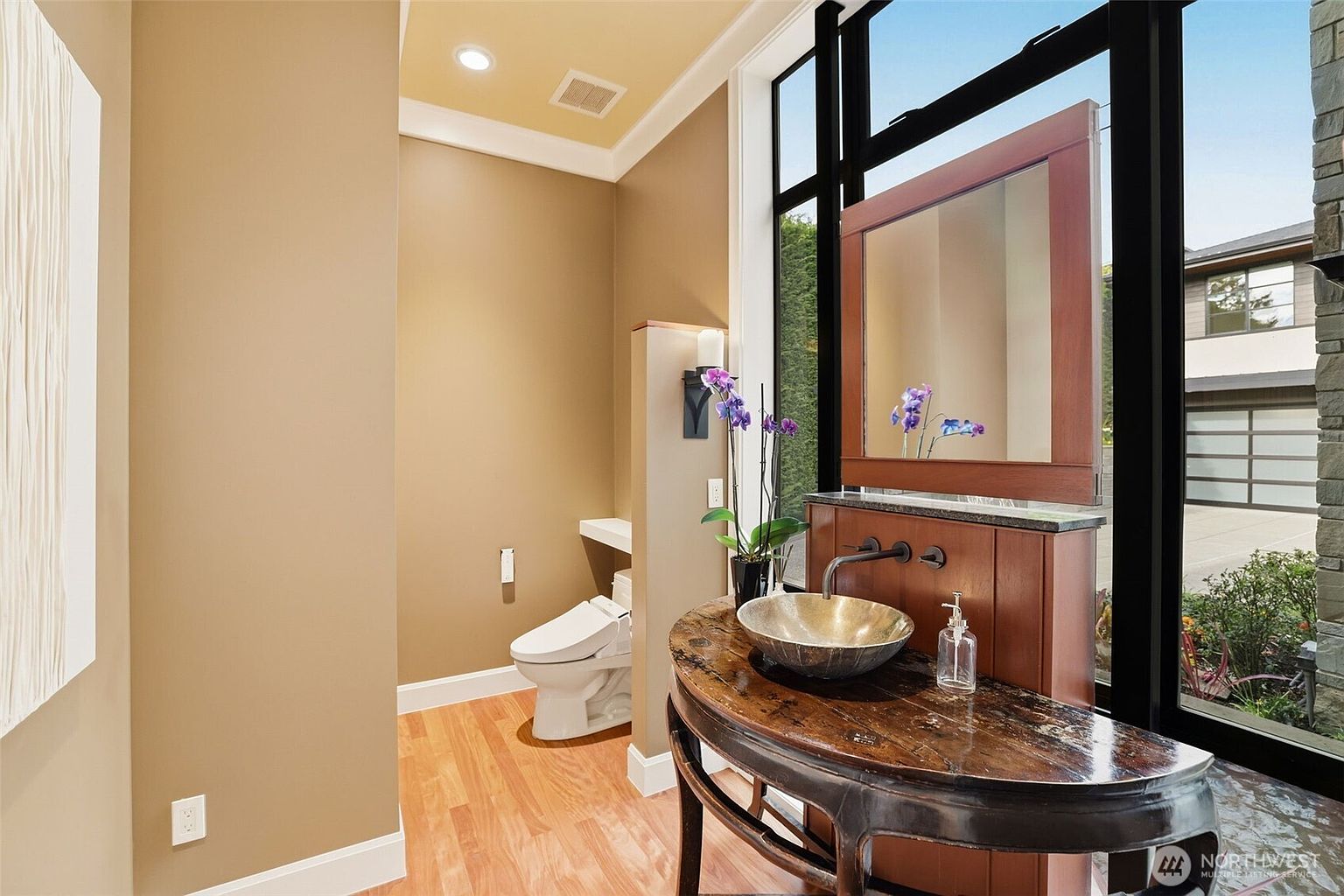 This unique powder room features a distinctive vanity with a dark, curved wooden base and a metallic vessel sink. A large mirror is suspended in front of tall windows that provide natural light and a view of the exterior, while a separate toilet area is tucked behind a partial wall. The space is finished with warm-toned walls and light wood flooring, creating a sophisticated and modern aesthetic.