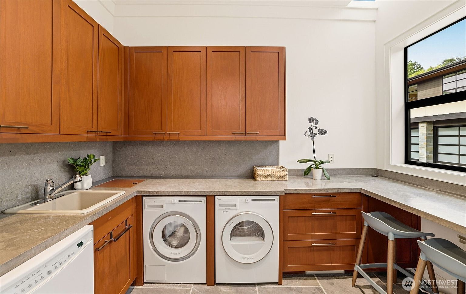 This functional laundry room features warm-toned wood cabinetry, a stone-look countertop, and a built-in sink. The space includes a side-by-side washer and dryer set, a small seating area with a stool, and a large window that allows for natural light. The overall aesthetic is clean, organized, and practical, blending utility with a polished design.
