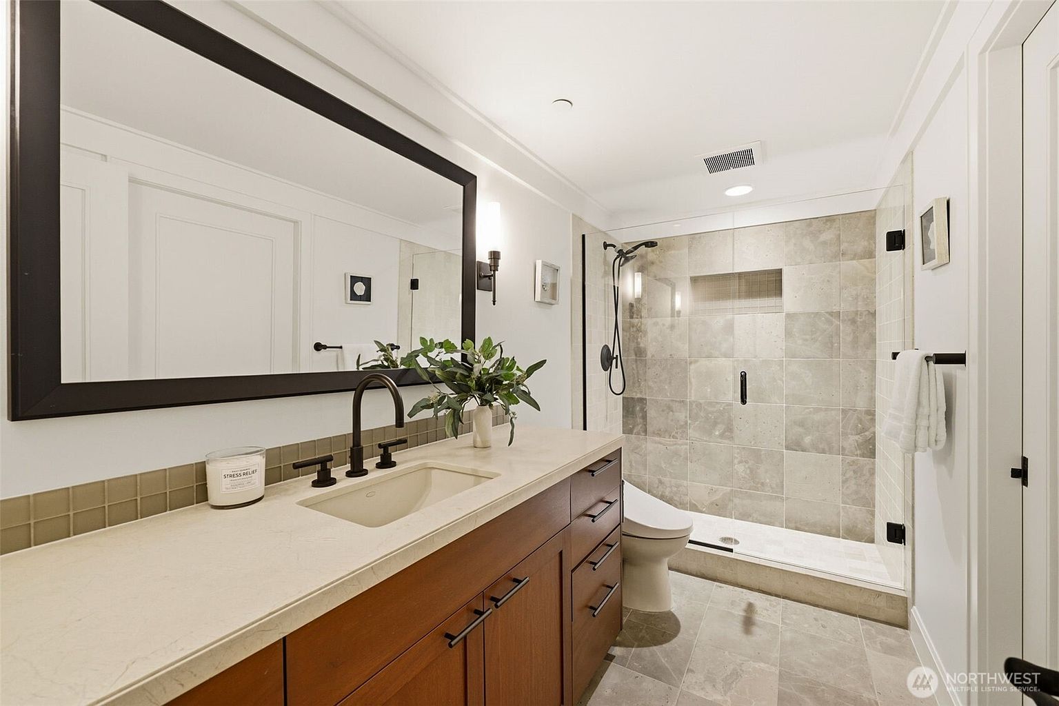 This modern bathroom features a sleek vanity with a light-toned countertop and dark wood cabinetry, complemented by a large mirror with a bold black frame. To the right, a walk-in shower is enclosed by glass and finished with neutral-toned square tiles, including a recessed niche for storage. The space is well-lit with recessed lighting and wall-mounted fixtures, creating a clean and contemporary atmosphere.