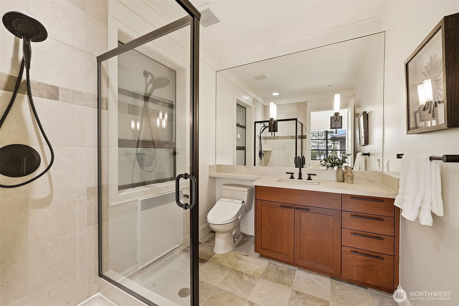 This modern bathroom features a glass-enclosed shower with a dark frame and a wall-mounted shower head. A wooden vanity with a stone countertop sits beneath a large, frameless mirror, complemented by a white toilet and neutral-toned floor tiles. The space is illuminated by sleek, vertical light fixtures, creating a clean and contemporary aesthetic.