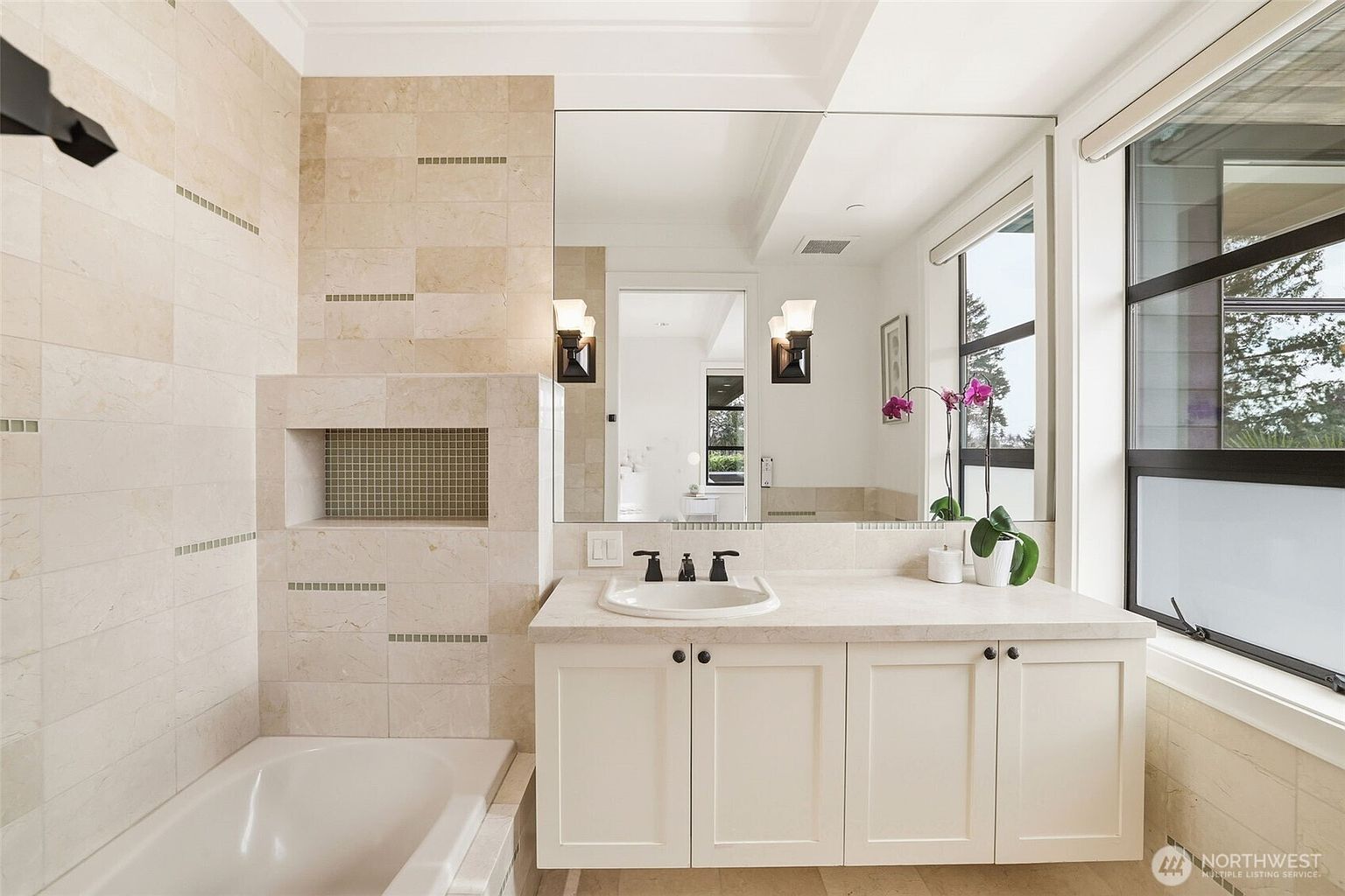 This bright and airy bathroom features a vanity with a light-toned stone countertop, a white undermount sink, and classic white cabinetry. A large, wall-mounted mirror reflects the space, while a tiled tub surround with a recessed niche adds a functional and decorative touch. Large windows to the right allow for ample natural light, creating a clean and inviting atmosphere.