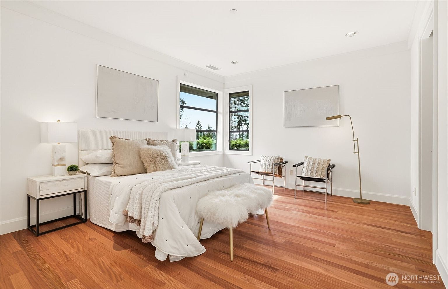 This bright and airy primary bedroom features warm-toned hardwood flooring and crisp white walls, creating a clean and inviting atmosphere. The room is furnished with a plush bed topped with textured linens, a fluffy bench at the foot, and two modern chairs near a corner window arrangement that provides ample natural light. The minimalist decor and neutral color palette emphasize the spaciousness and contemporary design of the suite.