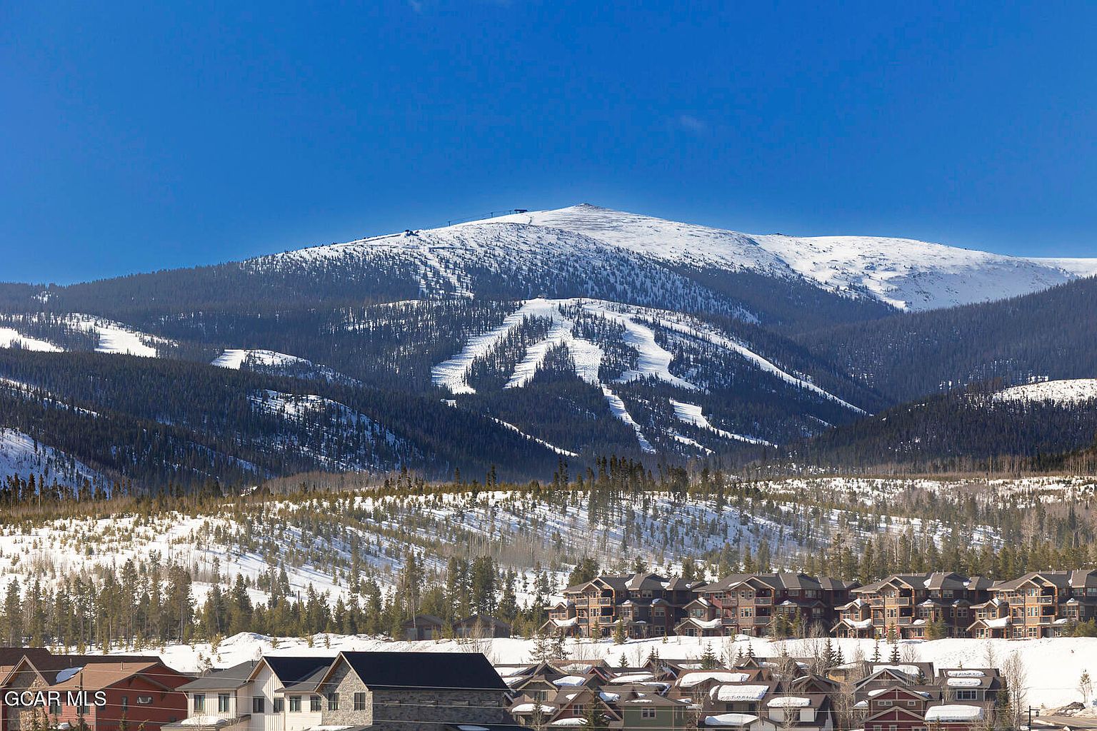 This image showcases a picturesque mountain view with a neighborhood of multi-story homes in the foreground. The homes feature a mix of architectural styles and colors, blending into the snowy landscape. The majestic, snow-covered mountain dominates the background, creating a stunning backdrop for the residential area.