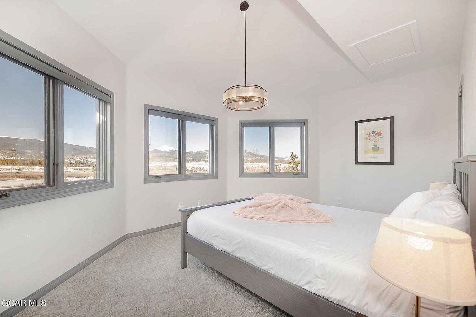 This is a bright and airy guest bedroom featuring a gray bed with white linens and a light pink throw. Three windows offer views of the surrounding landscape, and a modern light fixture hangs from the ceiling. The room is painted in neutral tones, creating a calming and inviting atmosphere.
