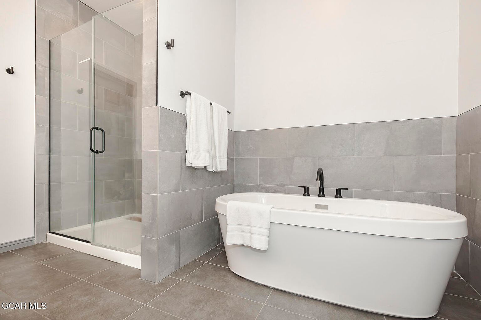 This is a well-lit primary bathroom featuring a modern freestanding bathtub with black fixtures and a white towel draped over the edge. The walls are partially tiled in gray, with the upper portion painted white. A glass-enclosed shower is visible to the left, and the floor is covered in gray tiles, creating a clean and contemporary aesthetic.