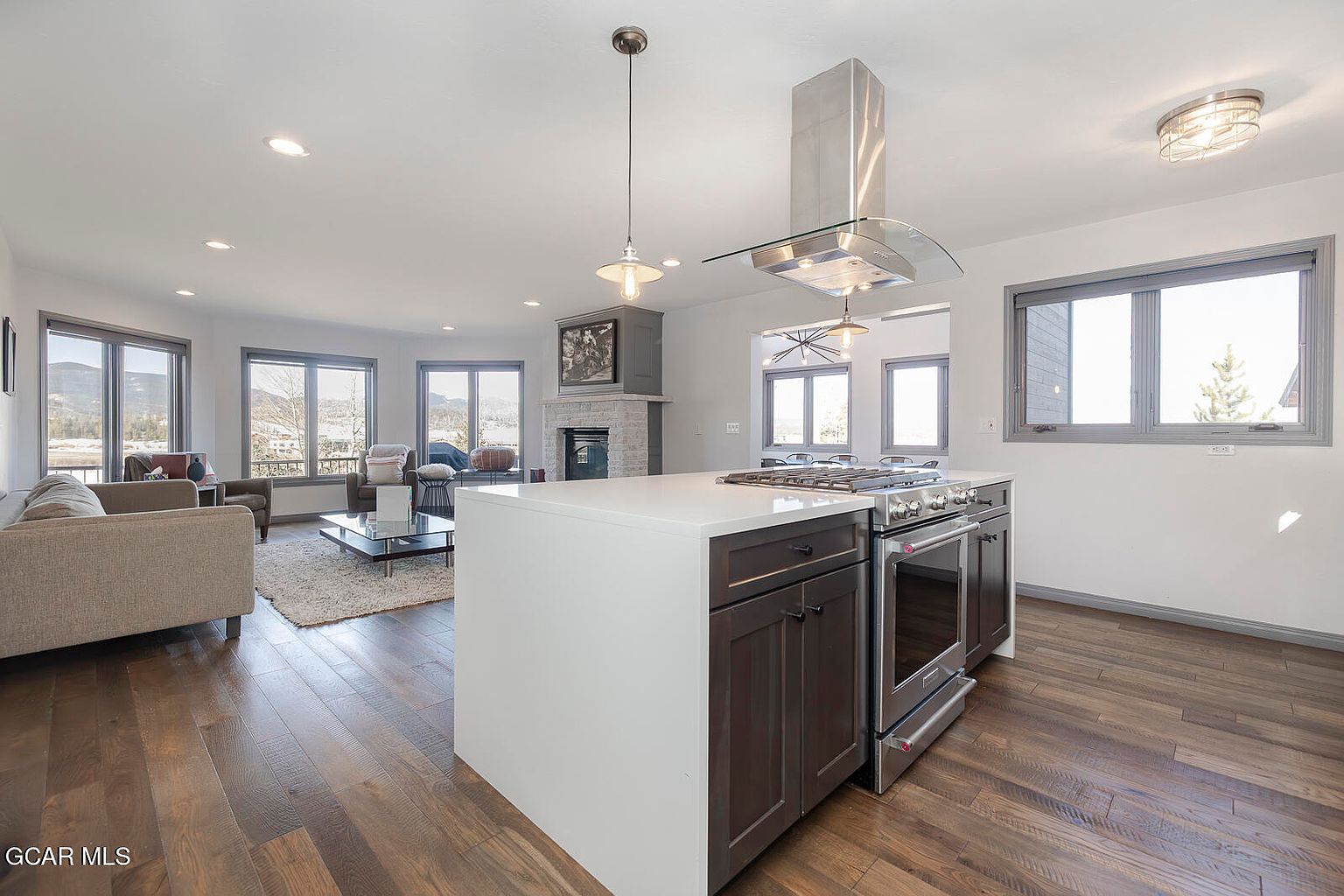 This interior shot showcases a modern kitchen seamlessly integrated with a living area. The kitchen features a central island with a white countertop and dark wood cabinetry, complemented by stainless steel appliances and a sleek range hood. The open floor plan allows for a spacious and inviting atmosphere, enhanced by natural light streaming through large windows.