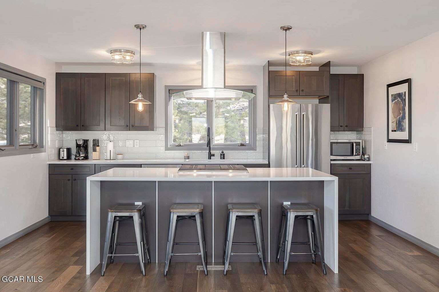 This is a well-lit kitchen featuring dark wood cabinets, stainless steel appliances, and a large island with a white countertop. The island has seating for four and is illuminated by pendant lights. A window above the sink provides natural light and a view of the outdoors, creating a warm and inviting atmosphere.