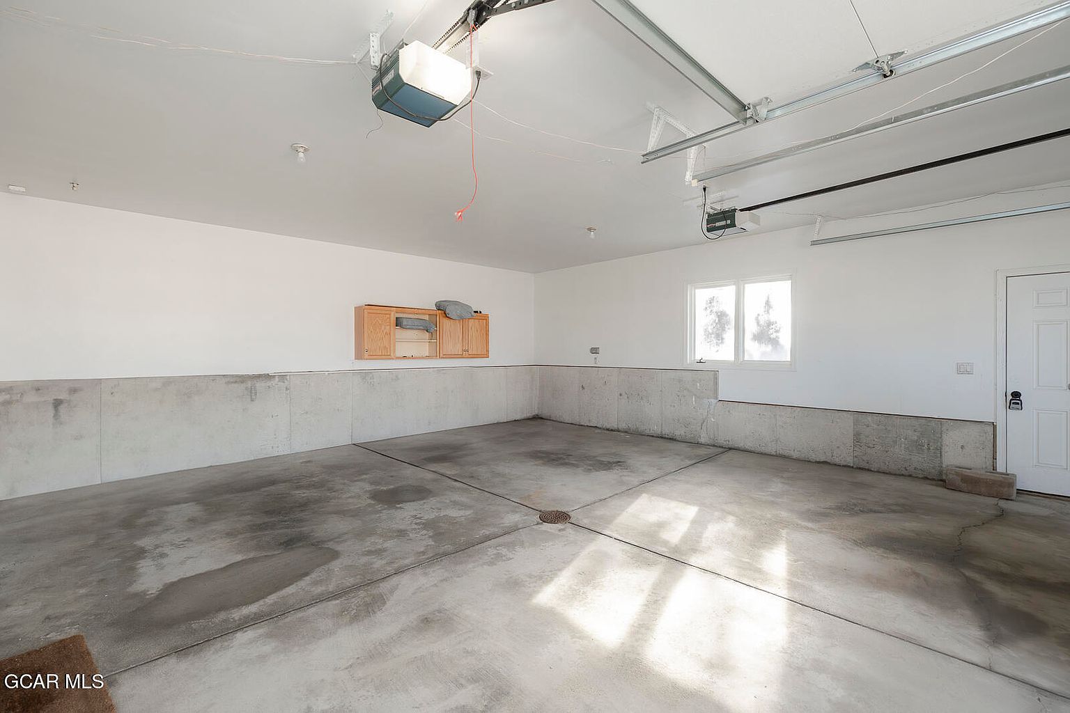 The image showcases a spacious and well-lit garage. The concrete floor is clean and expansive, complemented by a neutral-toned lower wall section. A wooden cabinet is mounted on the wall, and a window provides natural light, enhancing the garage's functionality and appeal.