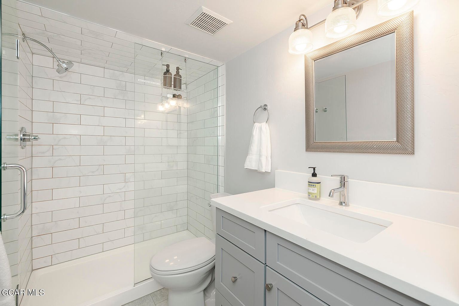 This is a well-lit bathroom featuring a glass-enclosed shower with white subway tile, a toilet, and a vanity with a gray cabinet and white countertop. A framed mirror hangs above the sink, complemented by modern lighting fixtures. The overall impression is clean and contemporary, making it an inviting space.