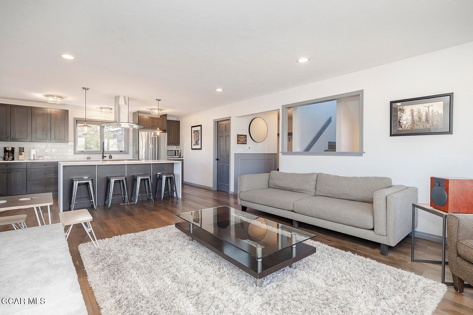 This is an interior shot of a living room and kitchen area. The living room features a gray sofa, a glass-topped coffee table on a light-colored rug, and hardwood floors. The kitchen has dark cabinets, stainless steel appliances, and a breakfast bar with stools, creating an open and inviting space.