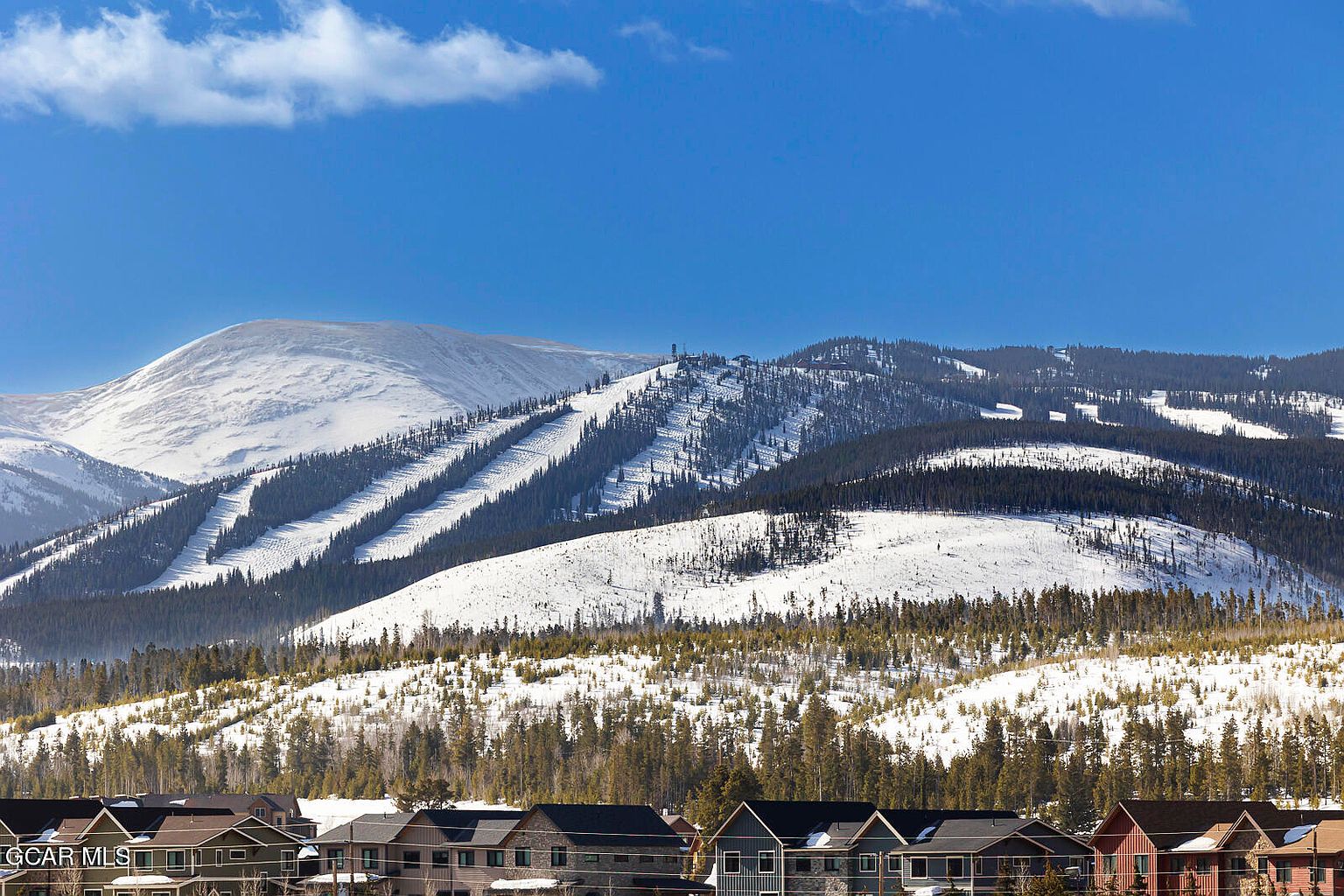 The image showcases a scenic view of residential buildings nestled in front of a snow-covered mountain range under a clear blue sky. The buildings appear to be multi-story townhouses or condos with varying rooflines and architectural styles. The snow-covered mountains in the background add a sense of grandeur and highlight the property's proximity to nature and recreational opportunities.