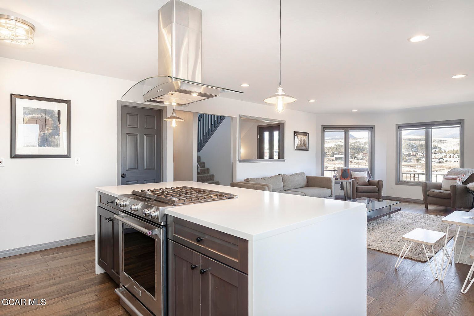 This interior shot showcases a modern kitchen that seamlessly transitions into a living area. The kitchen features a central island with a built-in stovetop and a sleek range hood, complemented by dark wood cabinetry and white countertops. The living area is bright and airy, with large windows offering scenic views and comfortable seating arrangements.