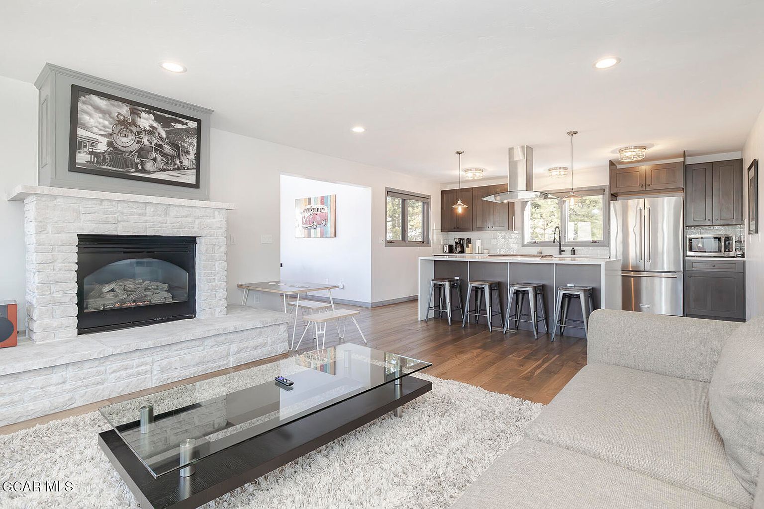 This is an interior shot of a living room that seamlessly blends into a modern kitchen. The living room features a stone fireplace with a framed picture above, a glass-top coffee table on a plush rug, and a comfortable-looking sofa. The kitchen boasts stainless steel appliances, dark cabinetry, a kitchen island with bar stools, and hardwood flooring, creating an open and inviting space.