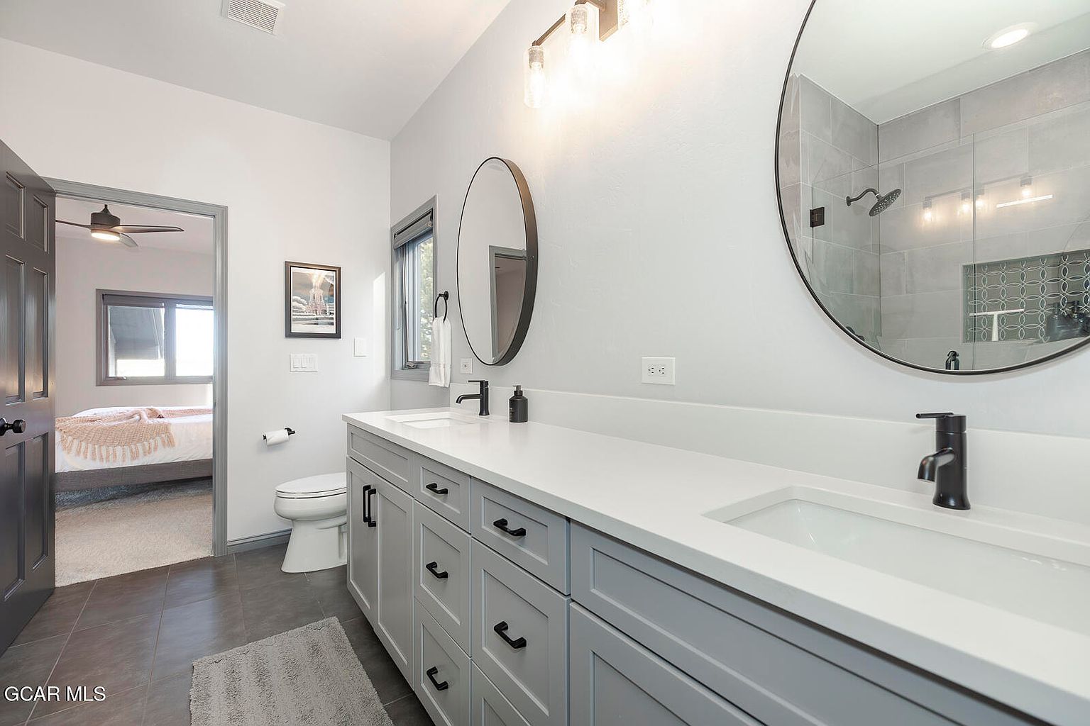 This is a well-lit primary bathroom featuring a double vanity with white countertops and gray cabinetry. Two round mirrors hang above the sinks, and modern black faucets add a touch of elegance. The bathroom has a clean and contemporary design, with a glimpse into an adjacent bedroom through an open doorway.