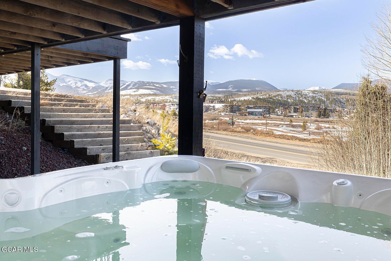 This image showcases a luxurious outdoor spa with a stunning mountain view. The spa is filled with clear water, inviting relaxation. The surrounding landscape includes a wooden deck, stairs, and a glimpse of the nearby town, creating a serene and picturesque setting.