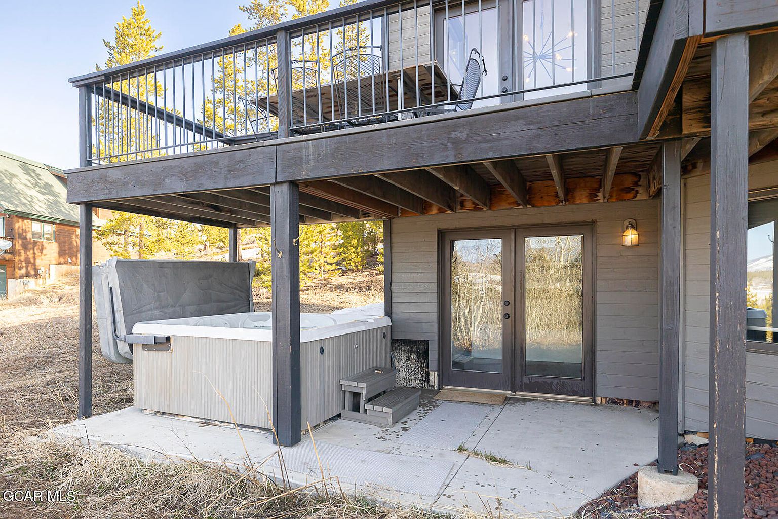 This image showcases a cozy outdoor patio area featuring a hot tub and a covered deck above. The hot tub is situated on a concrete slab, with steps leading up to it, and the deck above offers a shaded space for relaxation. The exterior of the building is clad in gray siding, creating a modern and inviting atmosphere.
