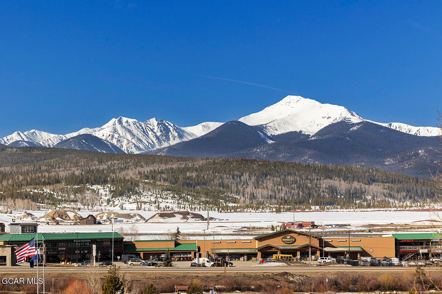 The image showcases a commercial building with a mountain backdrop under a clear blue sky. The building appears to be a retail complex, featuring multiple storefronts and a prominent sign for "Murdoch's". The surrounding landscape includes a mix of trees and snow-covered ground, suggesting a mountain town setting, ideal for attracting customers with scenic views.
