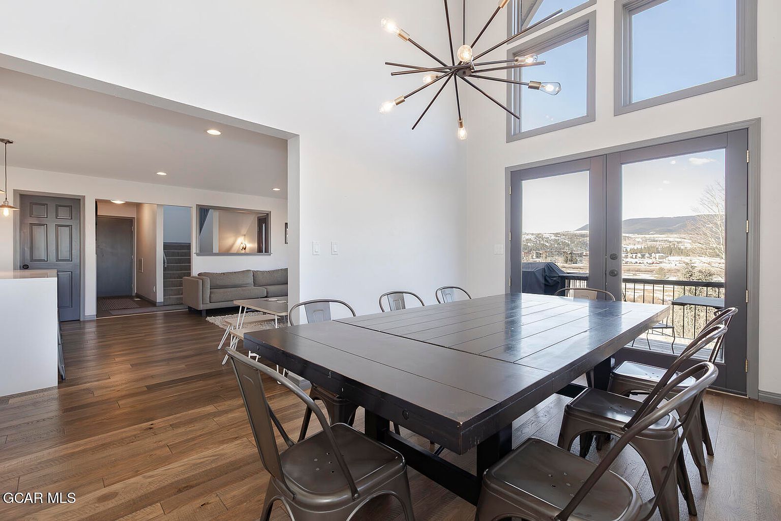 This interior shot showcases a dining room with a large, dark wood table surrounded by metal chairs. A modern chandelier hangs above the table, and large windows and a door offer a view of the landscape outside. The room has hardwood floors and a neutral color palette, creating a sophisticated and inviting atmosphere.