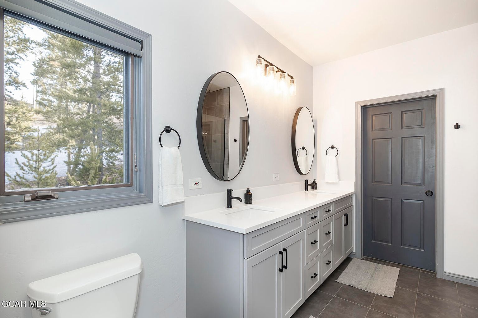 This is a well-lit primary bathroom featuring a double vanity with white countertops and gray cabinetry. Two oval mirrors hang above the sinks, complemented by a modern light fixture. A window provides natural light and a view of the outdoors, while a gray door adds a touch of sophistication.