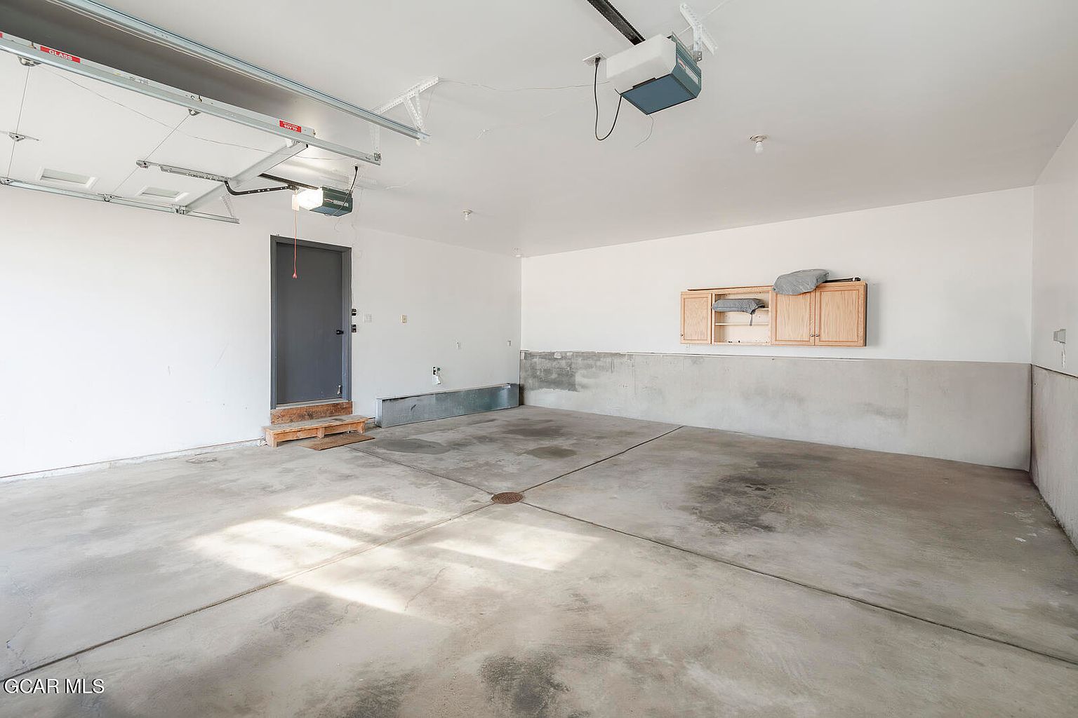 The image showcases an empty garage with a concrete floor and white walls. A gray door is visible on the left, and wooden cabinets are mounted on the back wall. The garage door mechanism is visible on the ceiling, and the space appears clean and spacious, ready for customization.