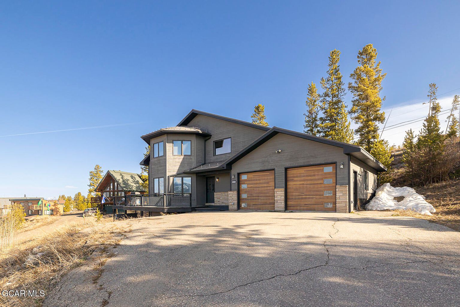 This is a front exterior view of a modern two-story home with a gray facade and wooden garage doors. The house features a unique architectural design with a rounded section and a deck. The driveway leads up to the house, and there are trees and some remaining snow visible in the background, suggesting a mountain or rural setting.