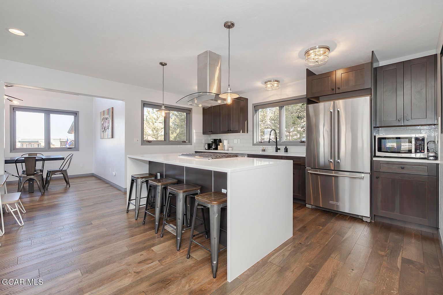 This is a well-lit kitchen featuring dark wood cabinets, stainless steel appliances, and a large white island with seating. The kitchen has a modern design with pendant lighting and a stainless steel range hood. The hardwood floors add warmth to the space, and the open layout connects to a dining area.