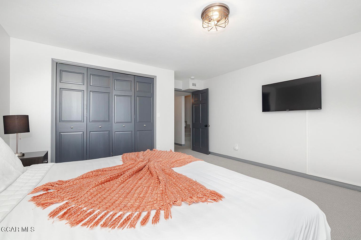 This is a bedroom featuring a bed with white linens and an orange throw blanket. There is a dark gray closet with paneled doors, a dark gray door leading to another room, and a flat screen television mounted on the wall. The room is painted in neutral tones, and there is a modern light fixture on the ceiling.