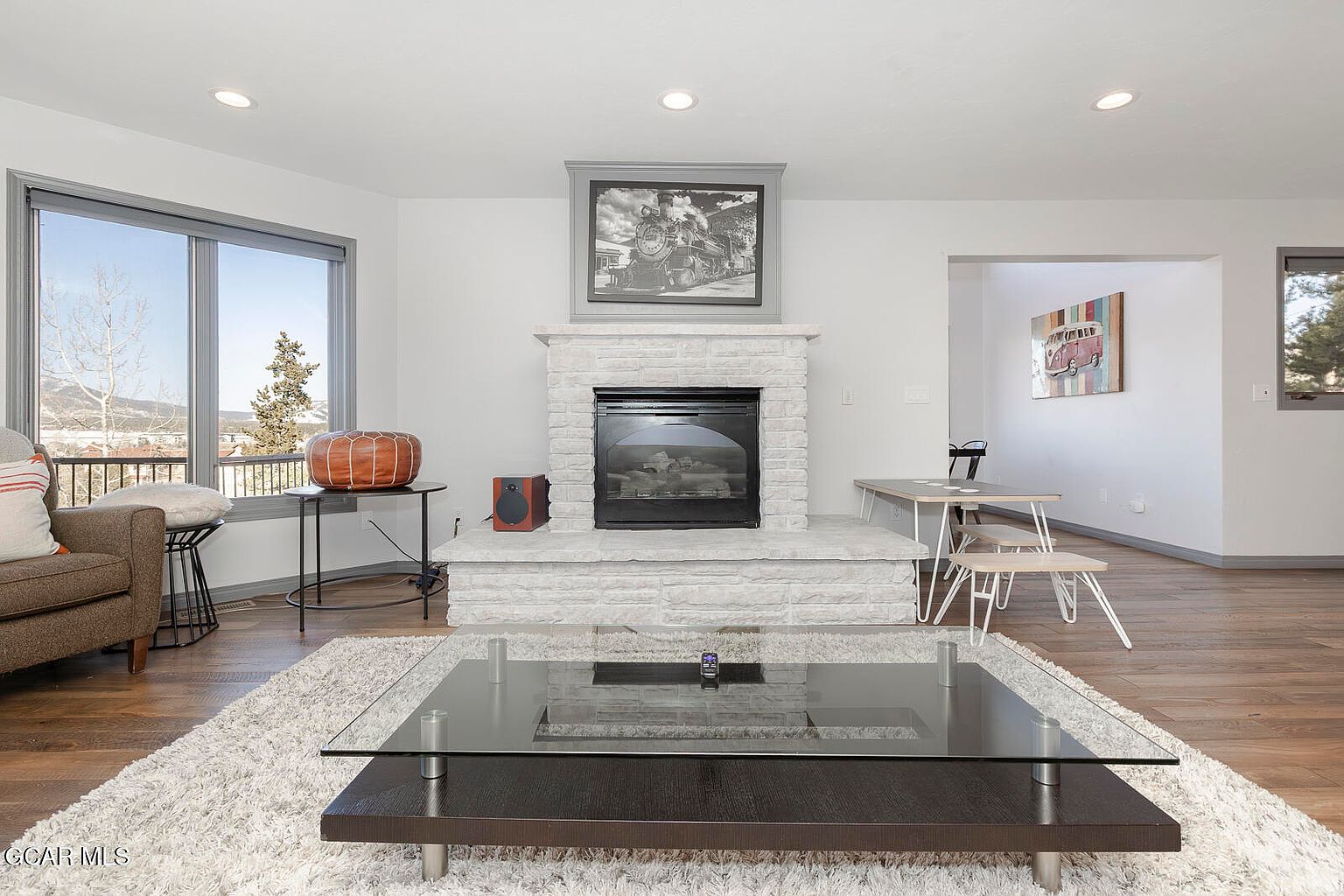 This is an interior shot of a living room featuring a fireplace with a black and white photo above it. The room has a modern aesthetic with a glass-top coffee table on a shag rug, a comfortable armchair, and a dining area in the background. Large windows provide natural light and a view of the outdoors.