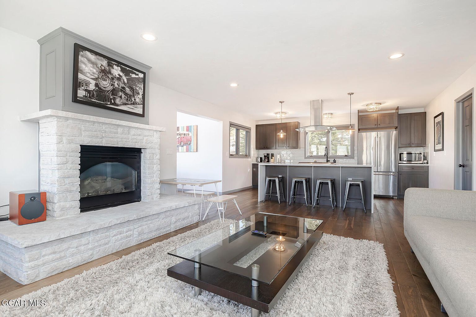 This is an interior shot of a living room that seamlessly blends into a modern kitchen. The living room features a stone fireplace with a black and white train picture above it, a plush rug, and a glass-topped coffee table. The kitchen boasts stainless steel appliances, dark cabinetry, and a breakfast bar with stools, creating an open and inviting space.