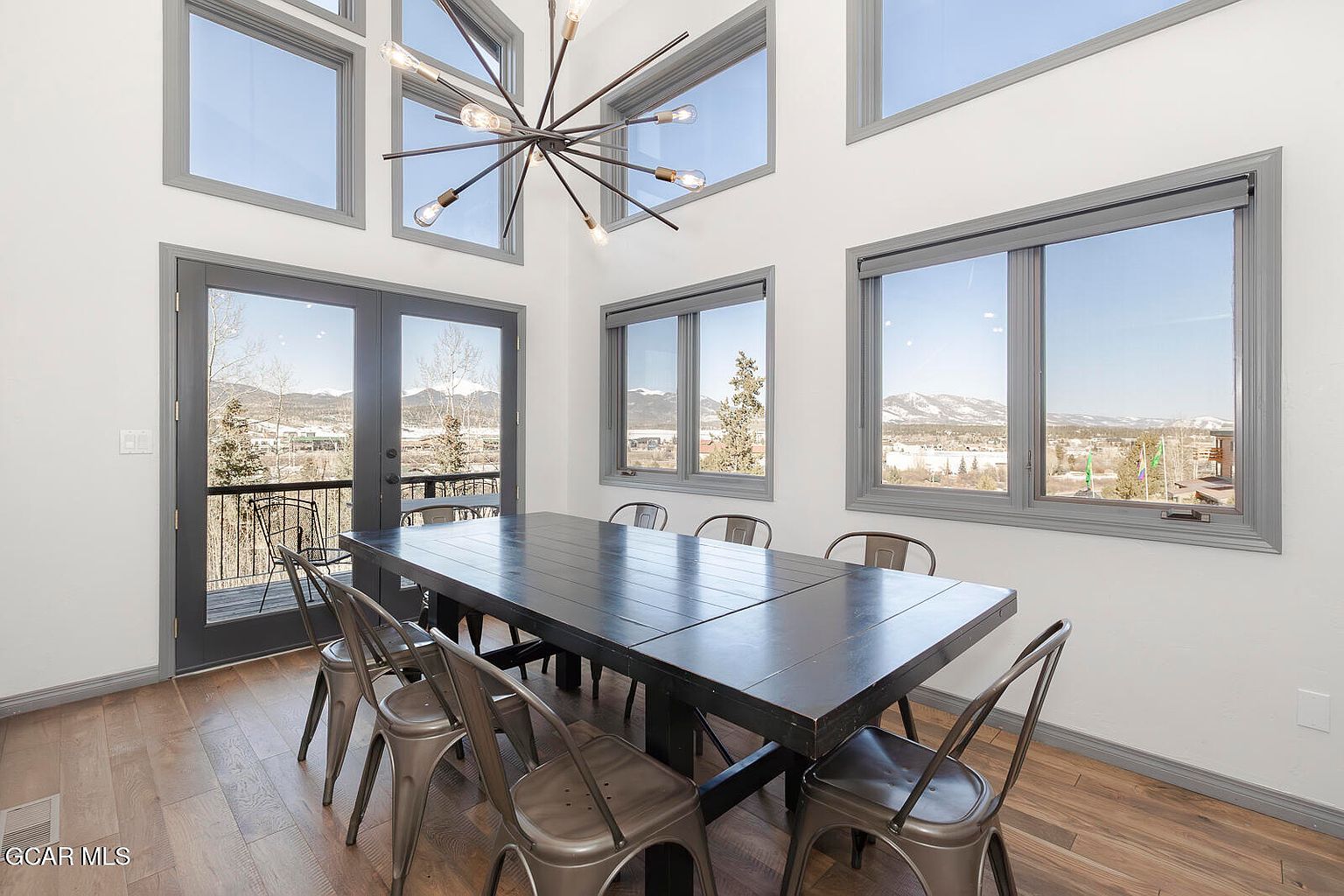 This is an interior shot of a dining room featuring a large, dark wood dining table surrounded by metal chairs. The room is well-lit with natural light streaming through multiple windows, offering a scenic view of mountains in the distance. An eye-catching, modern chandelier hangs above the table, adding a touch of contemporary style to the space.