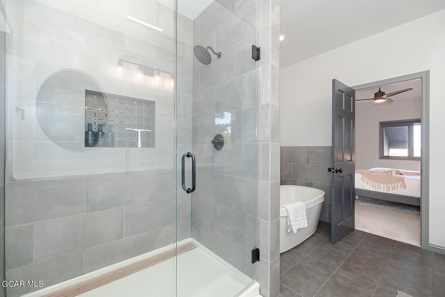 This is a well-lit primary bathroom featuring a modern walk-in shower with gray tiling and a glass door. A freestanding bathtub is visible next to the shower, and an open doorway leads into the bedroom. The bathroom has a clean and contemporary design.