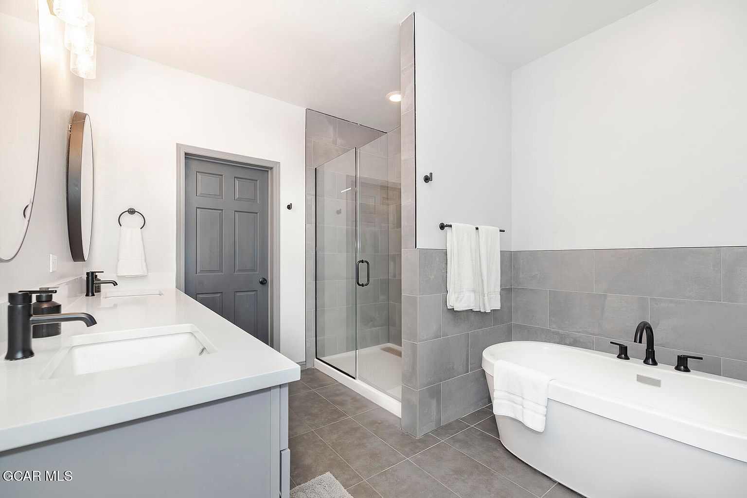 This is a well-lit primary bathroom featuring a double vanity with white countertops and gray cabinets, a glass-enclosed shower, and a freestanding bathtub. The walls are painted white with a gray tiled accent wall behind the bathtub, and the floor is covered in gray tiles. The fixtures are matte black, creating a modern and sophisticated look.