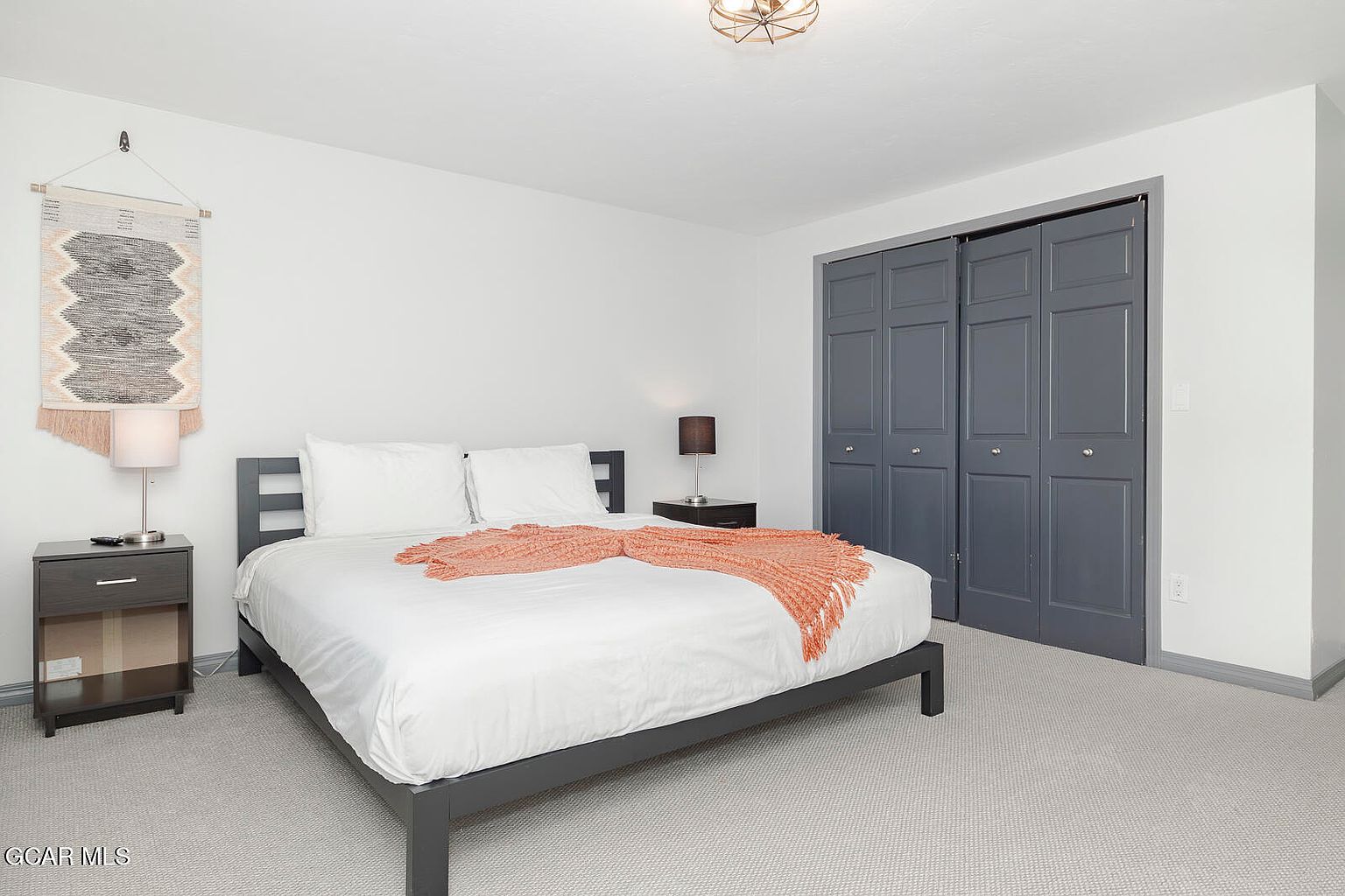 This is a well-lit primary bedroom featuring a queen-sized bed with white linens and an orange throw blanket. The room has a minimalist design with gray-toned furniture, including a nightstand and bed frame, complemented by a woven wall hanging. A closet with gray doors is visible in the background, and the room is carpeted in a neutral tone, creating a serene and inviting atmosphere.