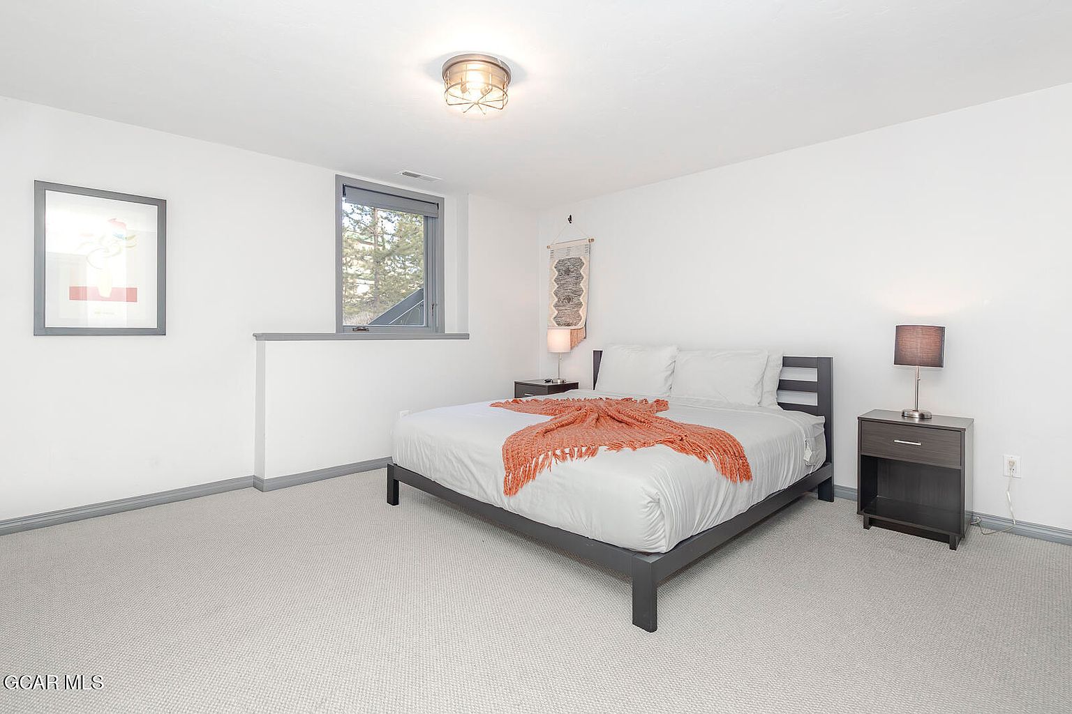 This is a bright and airy bedroom featuring a bed with white linens and an orange throw, flanked by two dark-colored nightstands. A framed picture hangs on the wall, and a window provides natural light. The room has a minimalist style with a neutral color palette, creating a calm and inviting atmosphere.