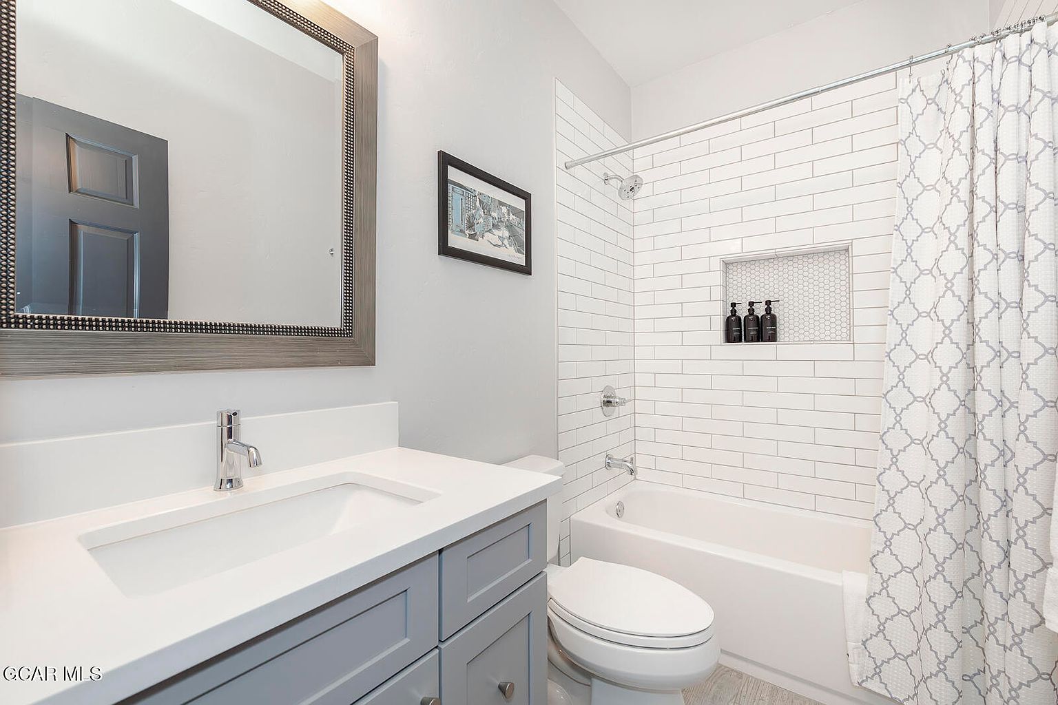This is a well-lit bathroom featuring a gray vanity with a white countertop and a rectangular sink. A framed mirror hangs above the vanity, reflecting a glimpse of a door. The bathroom includes a toilet and a bathtub with white subway tile surround and a shower curtain with a geometric pattern, creating a clean and modern aesthetic.