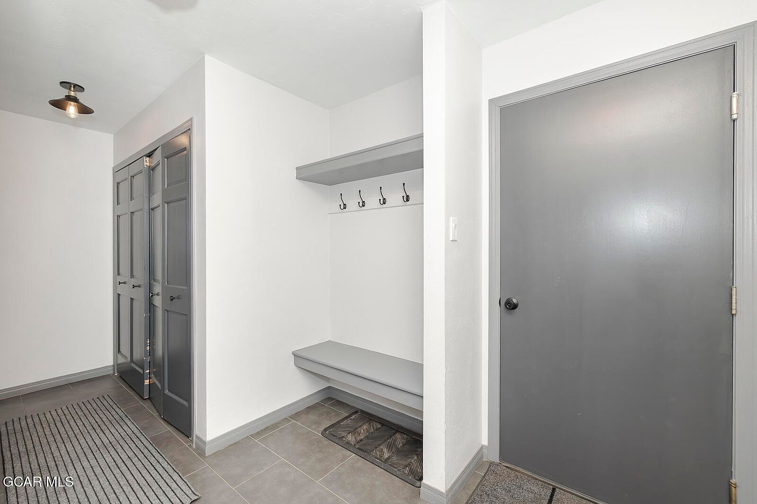 The image showcases an interior hallway featuring a built-in coat rack with a shelf and hooks, a gray bench, and a gray door. A closet with gray doors is visible on the left, and the floor is tiled in a neutral tone. The walls are painted white, creating a clean and modern aesthetic.