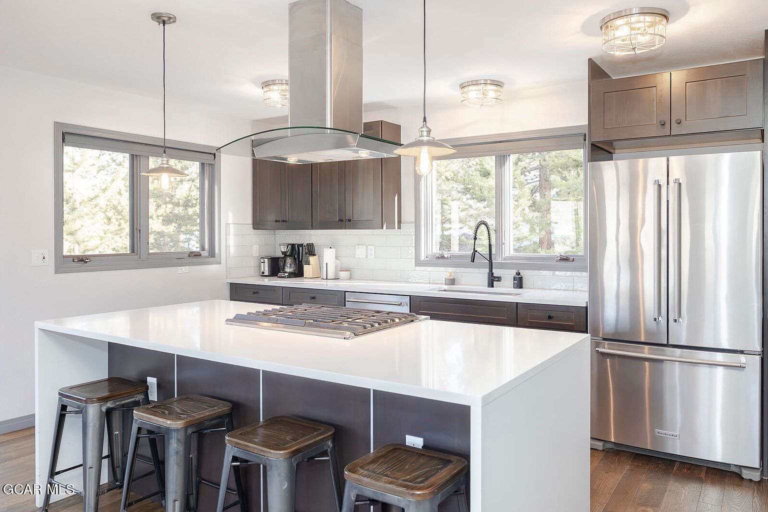 This is a well-lit kitchen featuring stainless steel appliances, dark wood cabinetry, and a large white countertop island with seating. The kitchen has a modern design with pendant lighting and a stainless steel range hood. Large windows provide natural light and views of the outdoors.
