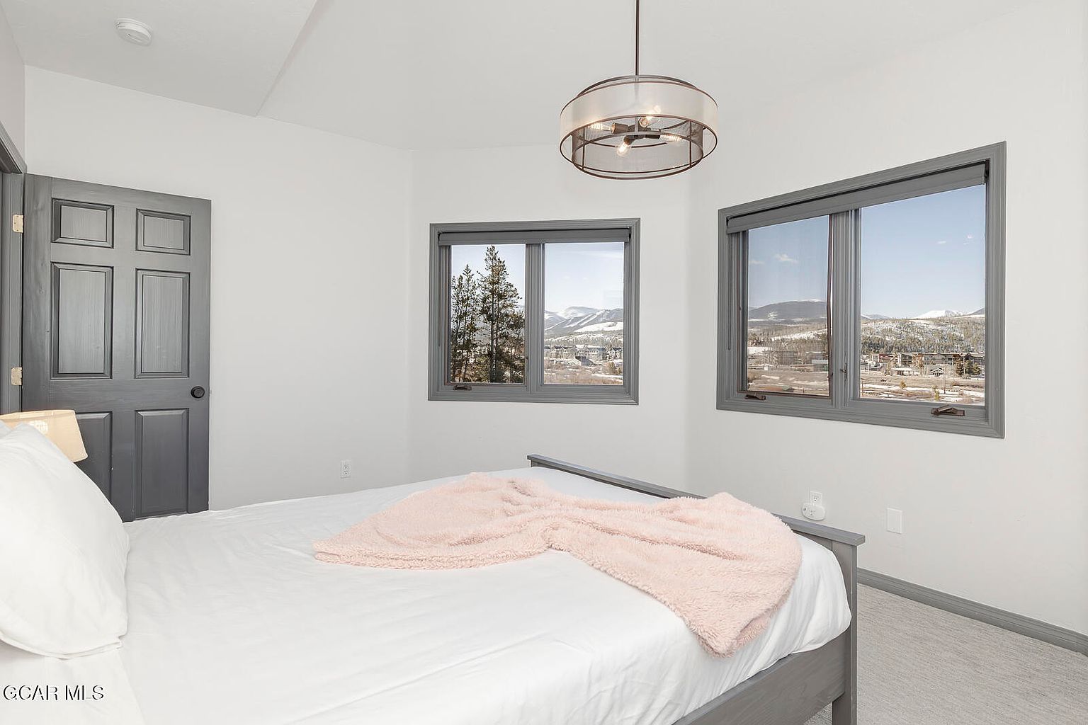 This is a bright and airy bedroom featuring a bed with white linens and a light pink throw blanket. The room has two windows offering scenic views of the mountains and surrounding landscape. A modern drum-style light fixture hangs from the ceiling, and the walls are painted a clean white, creating a serene and inviting atmosphere.