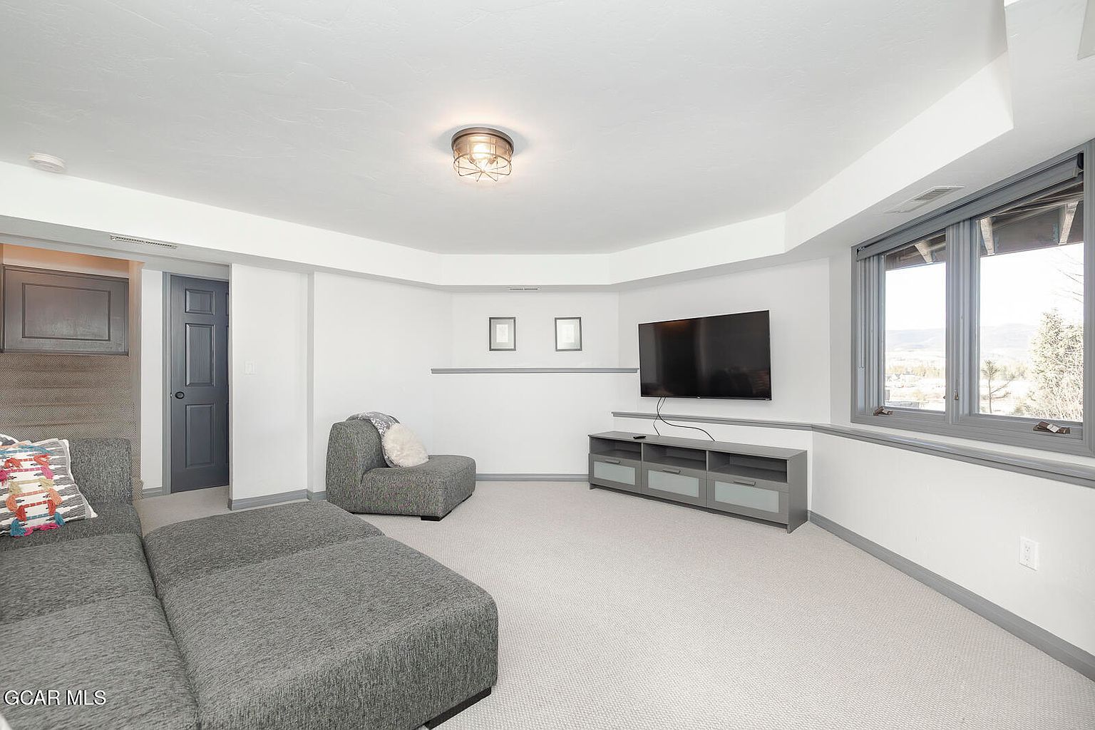 This is an interior shot of a living room featuring a gray sectional sofa, a matching armchair, and a gray media console beneath a wall-mounted television. The room has neutral-toned walls with gray trim, a light-colored carpet, and a window offering a view of the outdoors. The space is well-lit and appears modern and comfortable.